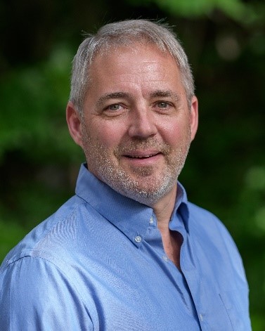 A white man with close cropped silver beard and hair, wearing a light blue button up shirt.